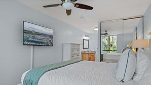Un dormitorio con cama, ventilador de techo, televisión montada en la pared, cómoda y un espejo grande.