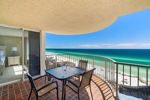A balcony with a table and chairs, overlooking a beach with clear turquoise water.