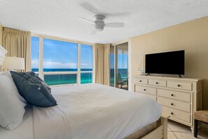 A hotel room with a large bed, a television, a ceiling fan, and a view of the ocean through large windows.