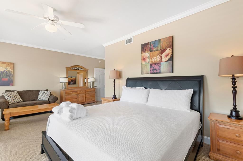 A bedroom with a large bed, a ceiling fan, a dresser, a nightstand, a lamp, a painting, and a couch.
