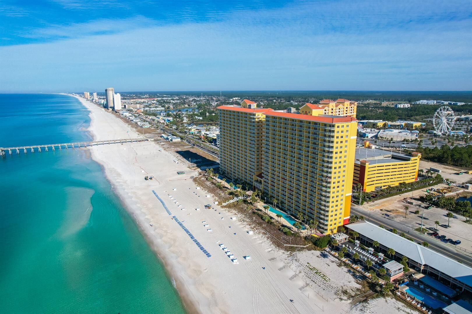 A coastal hotel with a beach view, a large parking lot, and a prominent Ferris wheel in the background.