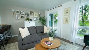 A living room with a grey sofa, a coffee table with a magazine, a white cabinet, and framed pictures on the wall. - Riverfront Oasis| Pool | King Bed Walk to Broadway (Nashville)