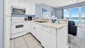 Une cuisine moderne avec des meubles blancs, un four à micro-ondes, un four et un lave-vaisselle. Une grande fenêtre offre une vue sur l’océan, et un balcon est agrémenté d’un canapé noir.