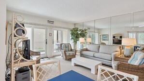 Amplia sala de estar con un sofá grande, una mesa de centro y un televisor sobre un mueble bajo. Hay un balcón con una silla y una mesa, y un espejo grande que refleja la habitación.