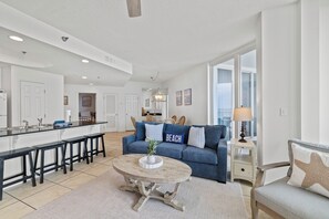 Een ruime woonkamer met keuken, een blauwe bank met een 'BEACH' kussen, een ronde salontafel en uitzicht naar buiten.