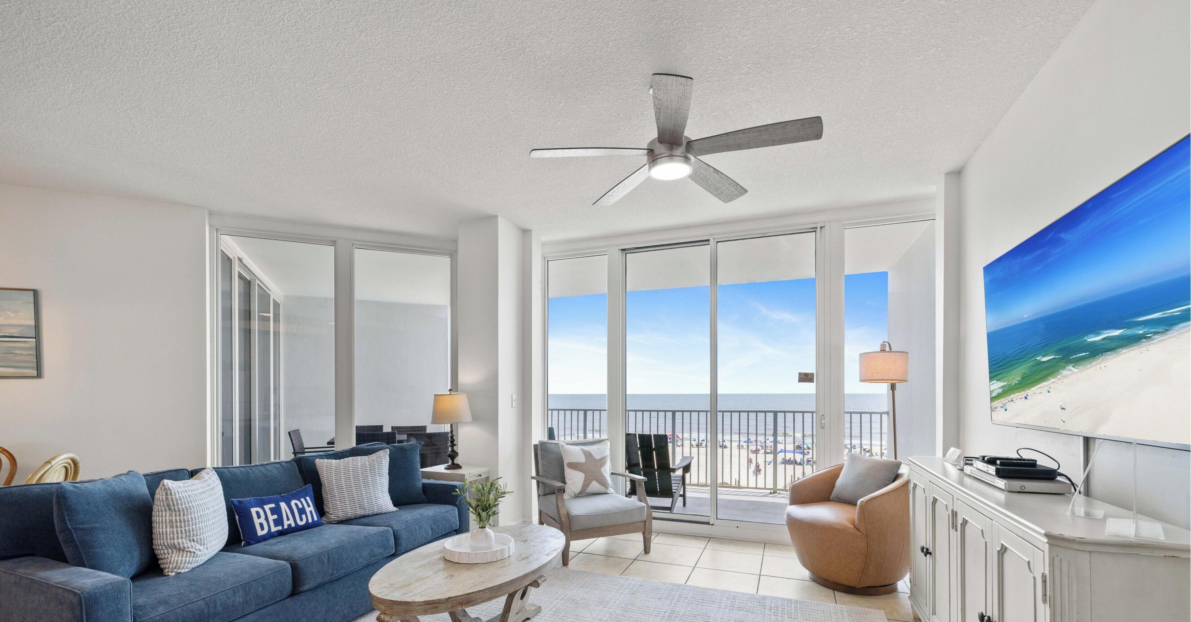 A spacious living room with a large window overlooking a beach, a ceiling fan, and a flat-screen TV on a cabinet.