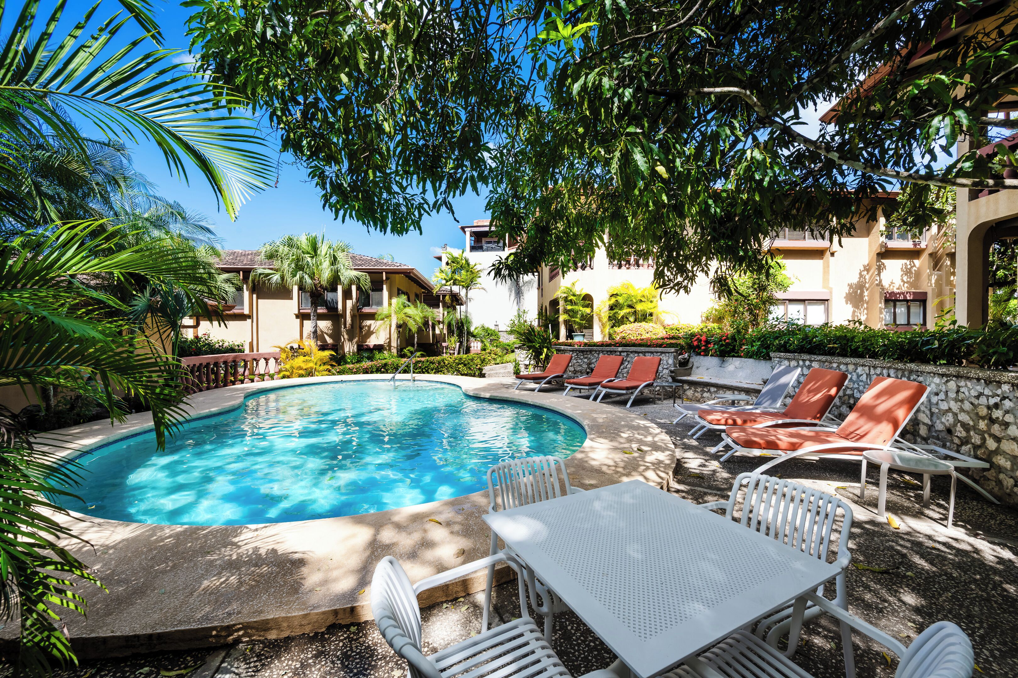 Uma piscina cercada por espreguiçadeiras e uma mesa, com um edifício ao fundo.