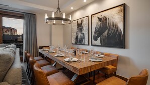 A dining area with a wooden table set for six, surrounded by leather chairs. There are two large horse paintings on the wall, a chandelier above the table, and a view of buildings through a window with curtains.