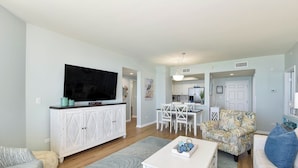 A living room with a TV, white furniture, a dining area, and a kitchen in the background. - Charming, bright beachfront condo w/private balcony! 🌴 (Destin)