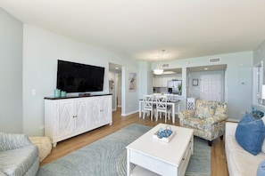 A living room with a TV, white furniture, a dining area, and a kitchen in the background.