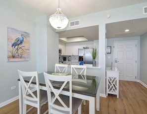 A dining area with a glass table and white chairs, a chandelier, a kitchen in the background, and a door with a sign.