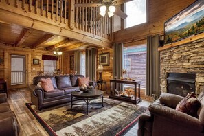 L’intérieur d’une cabane en rondins est agrémenté d’une cheminée, de poutres en bois au plafond, d’un canapé en cuir et d’un manteau de cheminée en pierre.