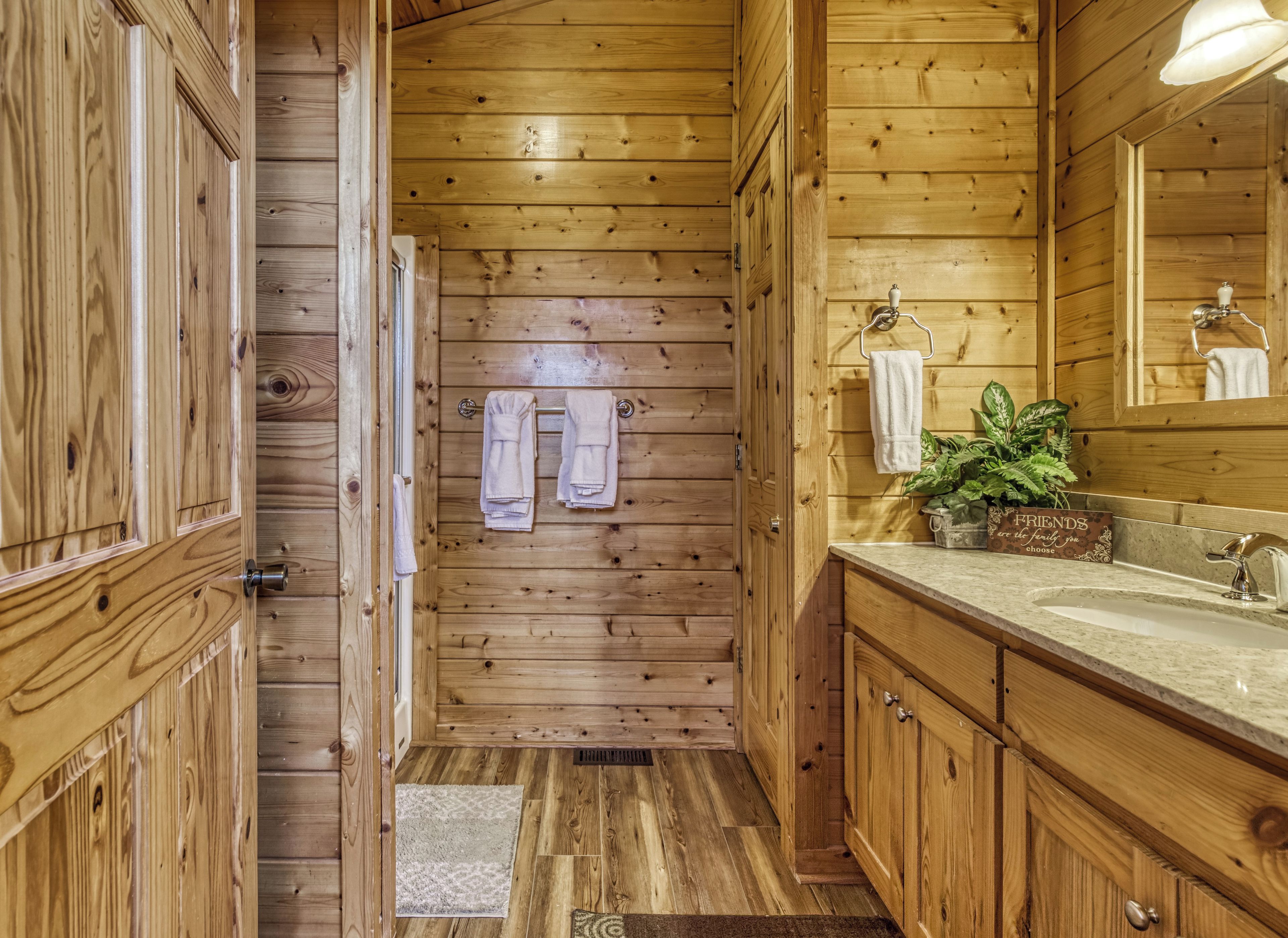 Ein Badezimmer einer Holzhütte mit Holztür, Handtuchhalter mit Handtüchern, Waschbecken, Spiegel und einer Pflanze auf der Ablagefläche.