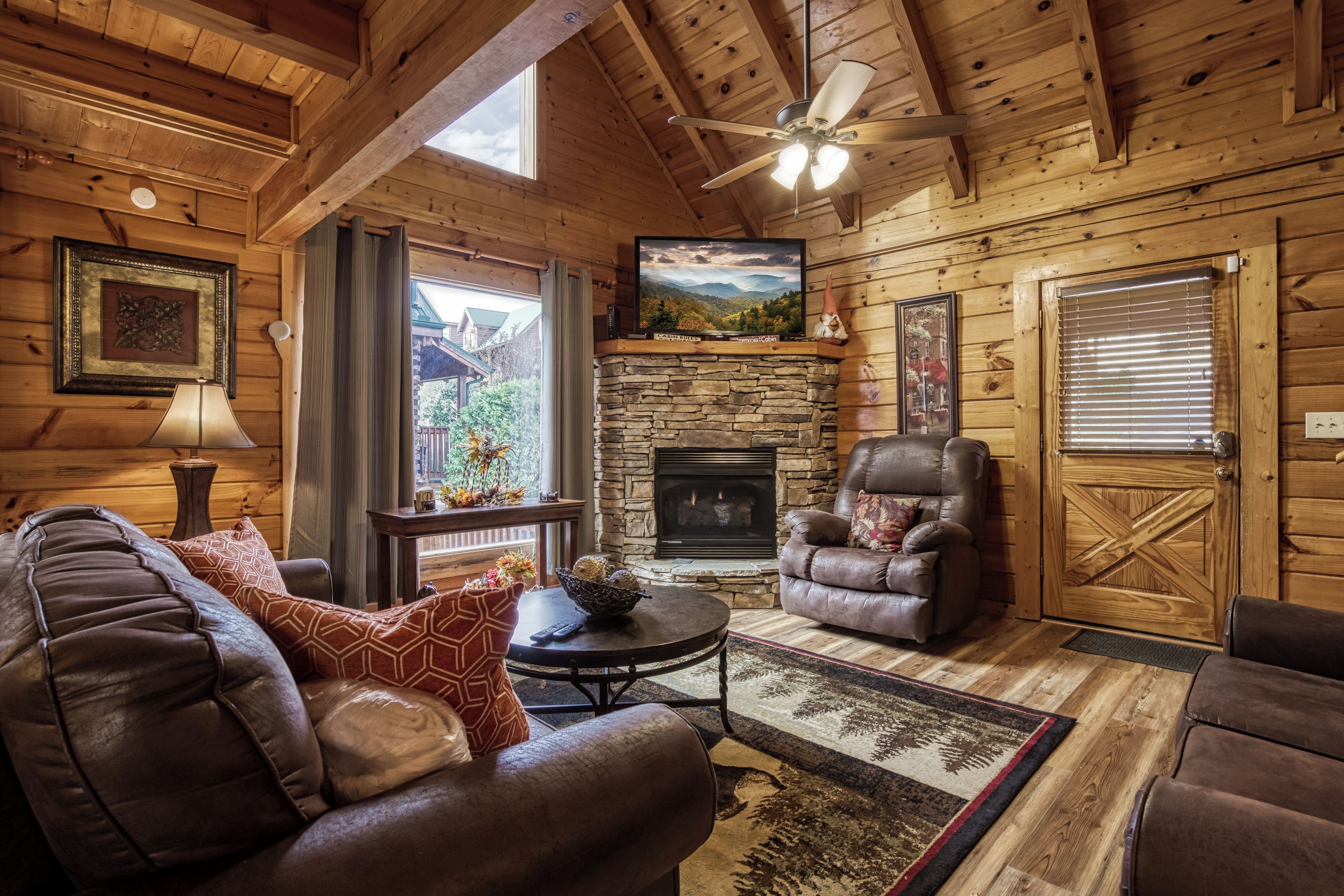 A cozy cabin interior with a stone fireplace, leather armchairs, a television, and a ceiling fan.
