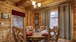 Une salle à manger en bois avec une table ronde dressée pour quatre personnes, des chaises en bois, un lustre et une fenêtre avec des rideaux.