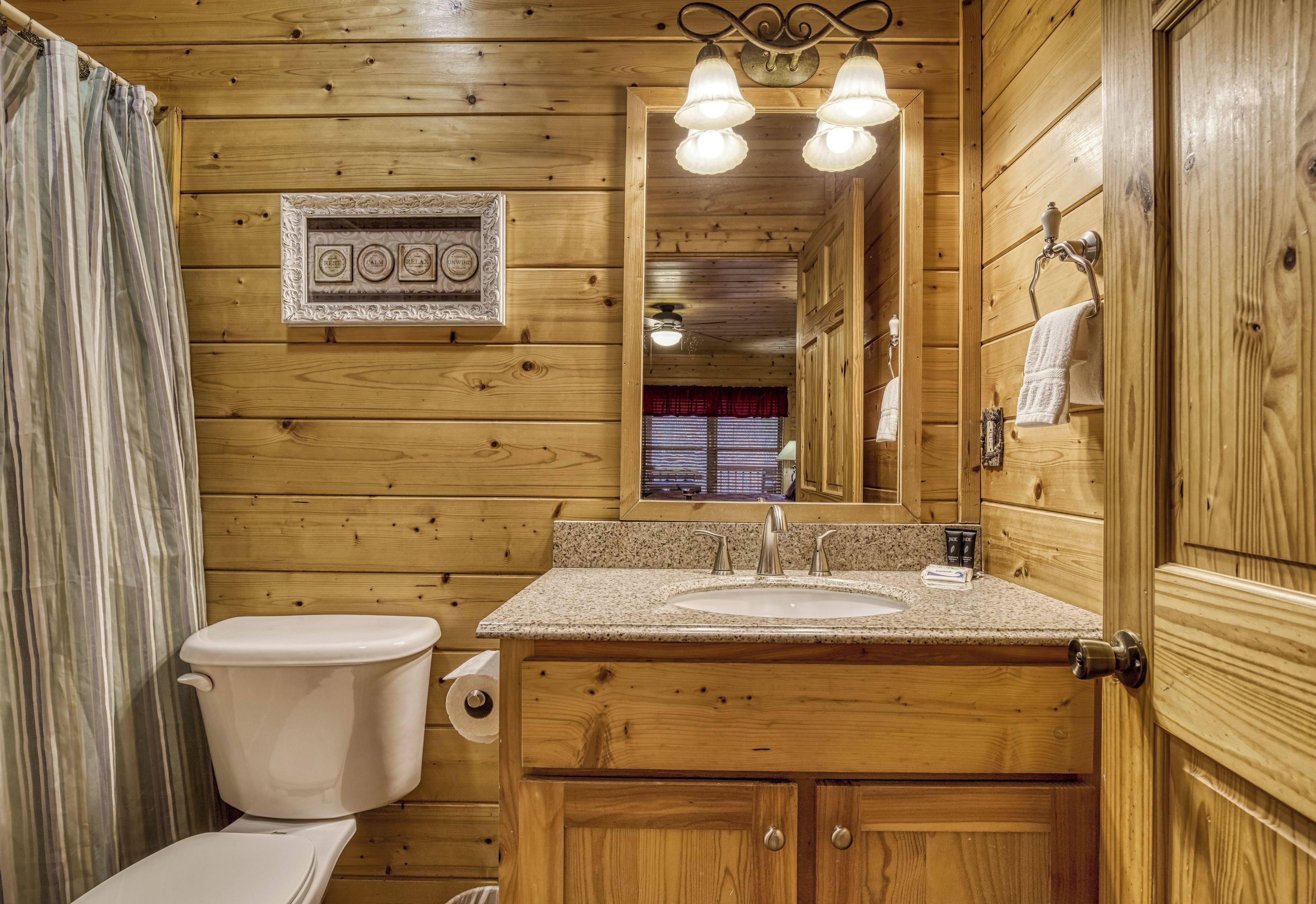 Un baño de madera con inodoro, lavabo y tocador. Hay un espejo sobre el lavabo y un cuadro en la pared.