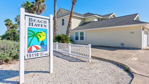 Un letrero con temática de playa para Beach Haven, una casa de dos pisos con garaje, rodeada por una cerca de piquetes blancos y palmeras.
