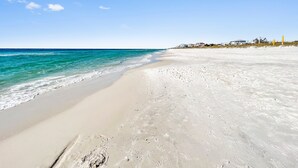 Una playa con agua azul cristalina, orilla arenosa y casas al fondo.