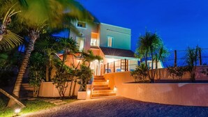 A modern two-story house with a well-lit entrance, a staircase, and a landscaped garden with palm trees. - Oceanfront Akumal Villa With Pool Near Snorkeling & Restaurants (Akumal)