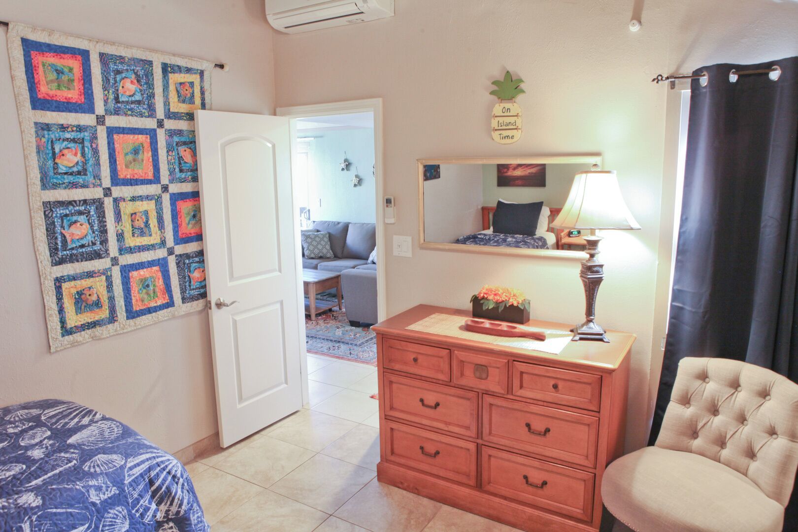 Una habitación con un tapiz acolchado en la pared, un aparador de madera con cajones, una silla, una lámpara y un espejo.