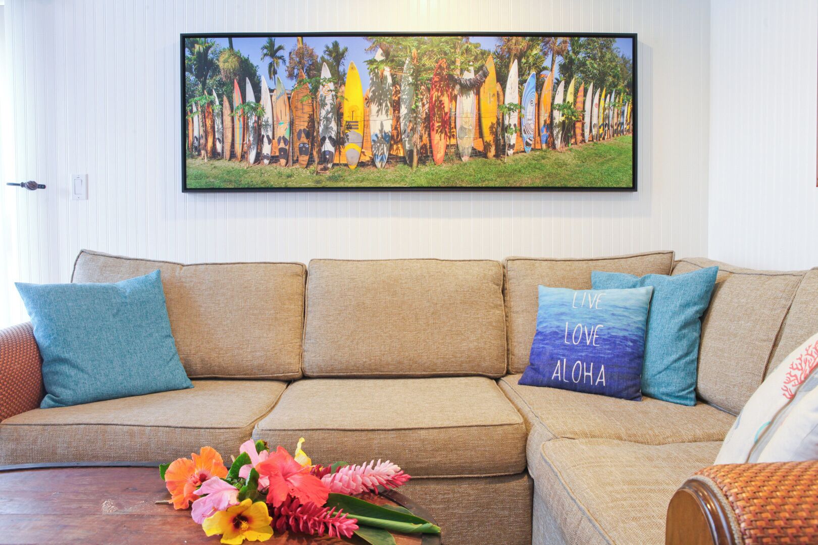 A beige sectional sofa with teal and blue cushions, a framed tropical surfboard mural, and a wooden coffee table with colorful flowers.