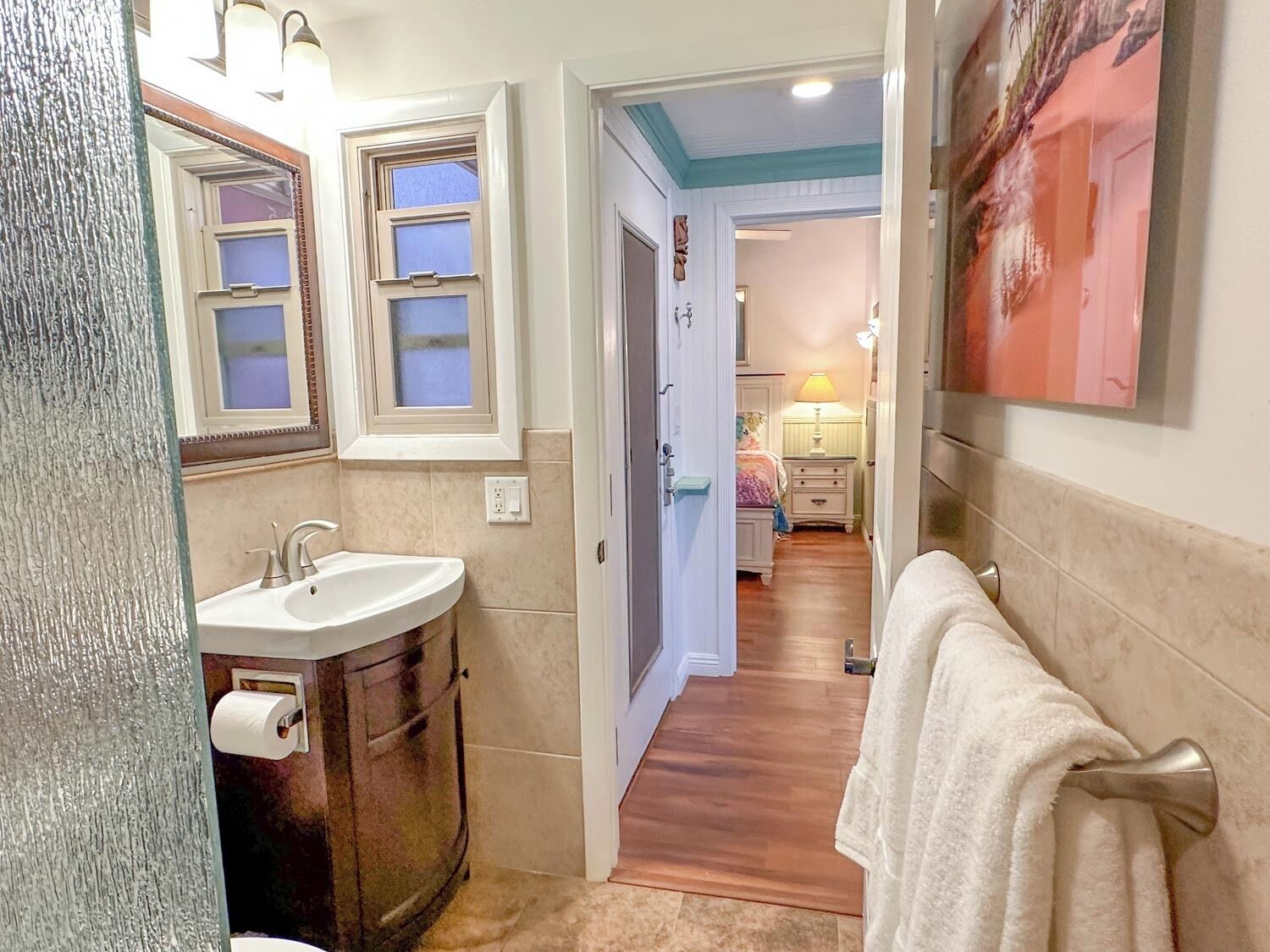 A narrow hallway leading to a bedroom with a wooden floor, a white sink, and a towel rack.