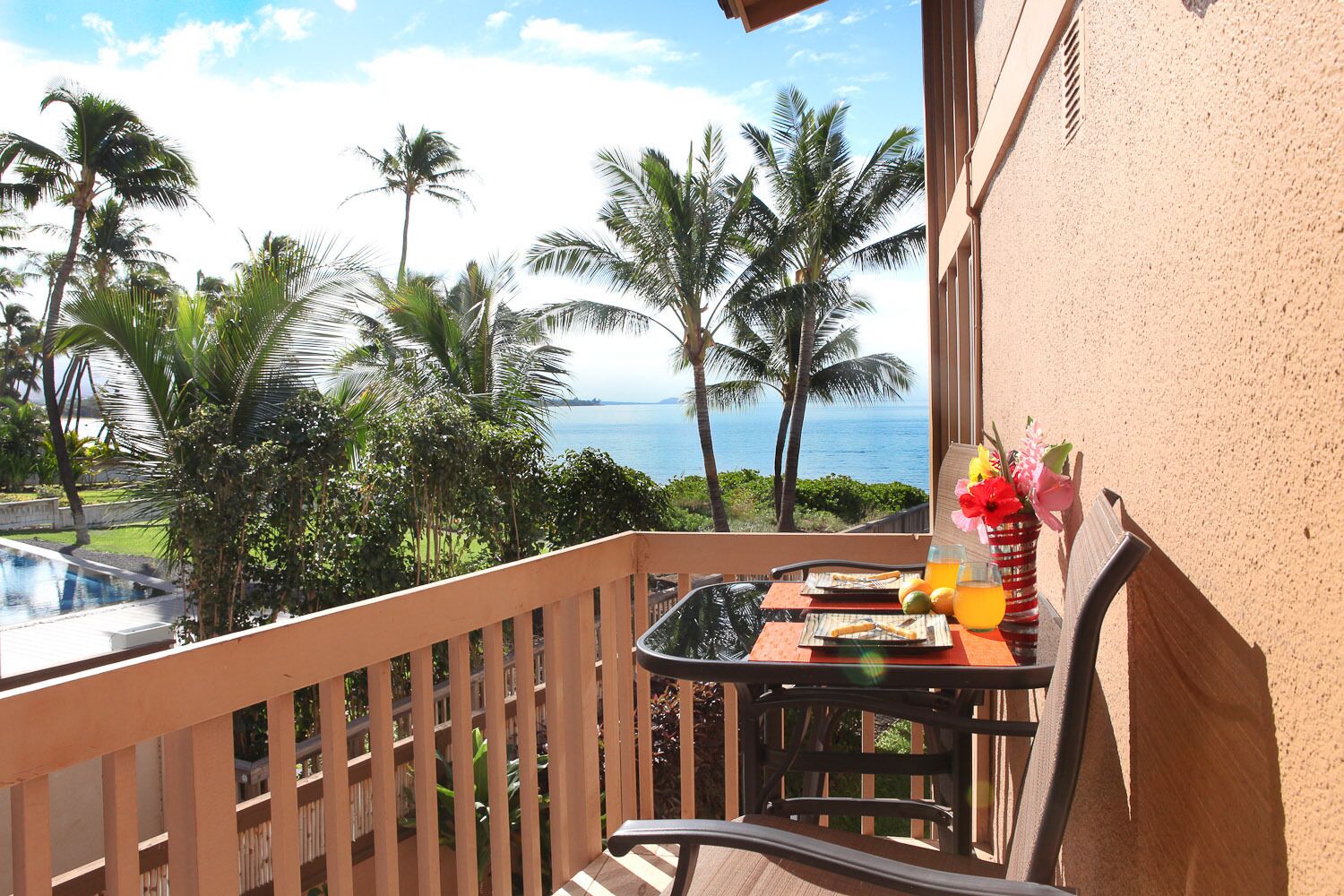 Un balcon avec une table dressée pour le petit-déjeuner, offrant une vue sur un paysage tropical avec des palmiers et l’océan.