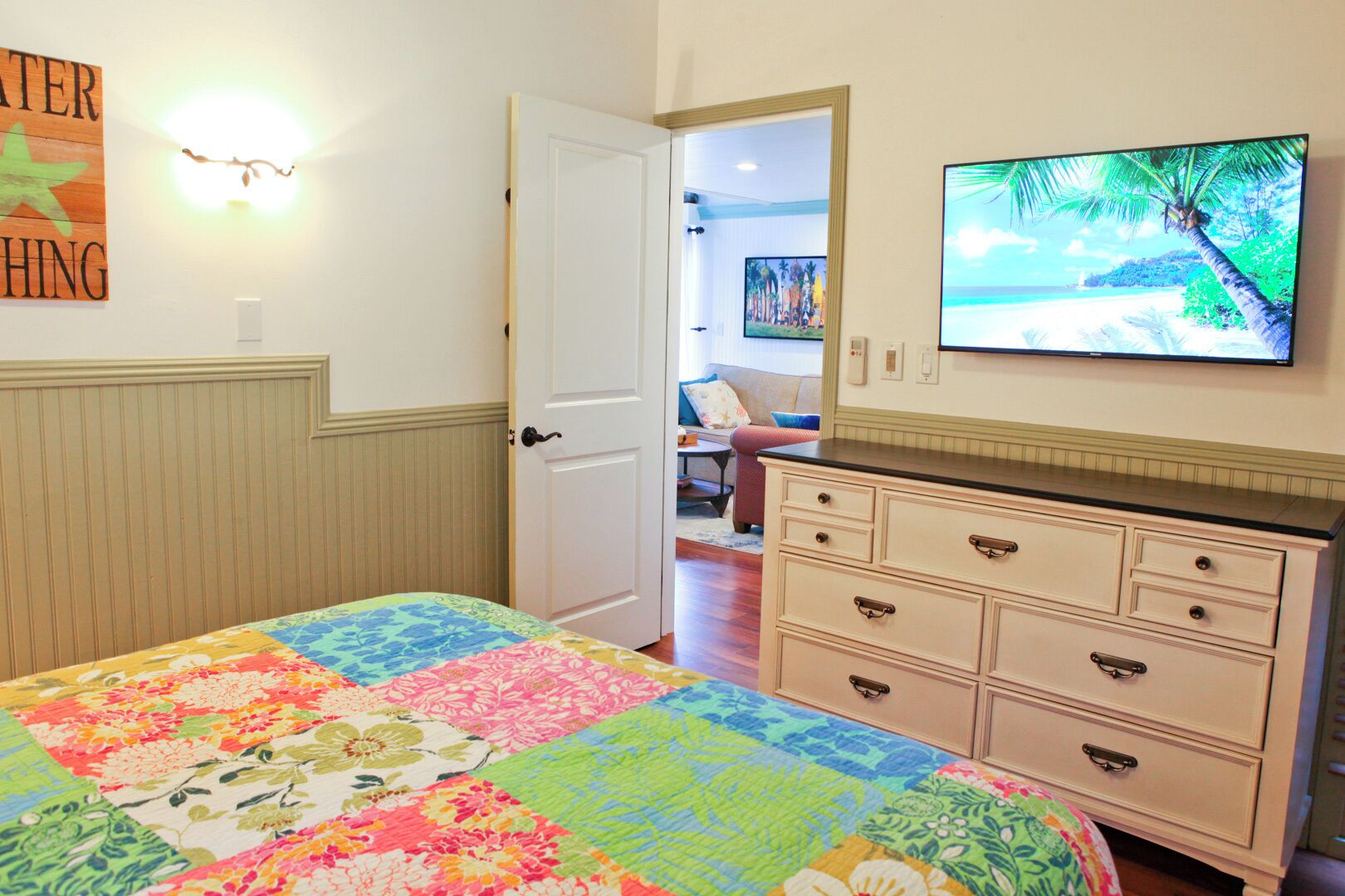A bedroom with a colorful quilt, a dresser, a television displaying a beach scene, and a door leading to another room.