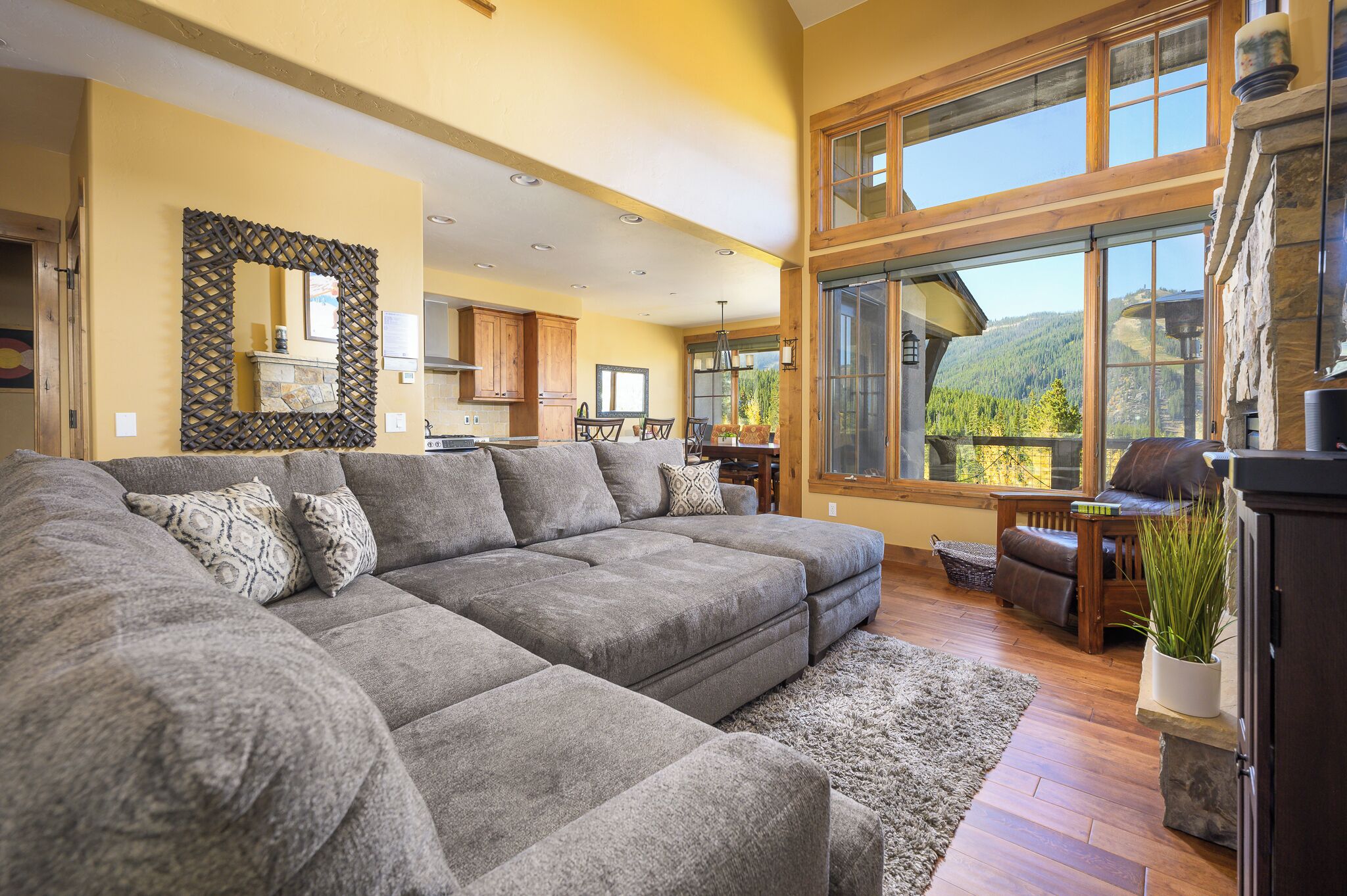 A spacious living room with a large sectional sofa, wooden flooring, and a view of a mountainous landscape through large windows.