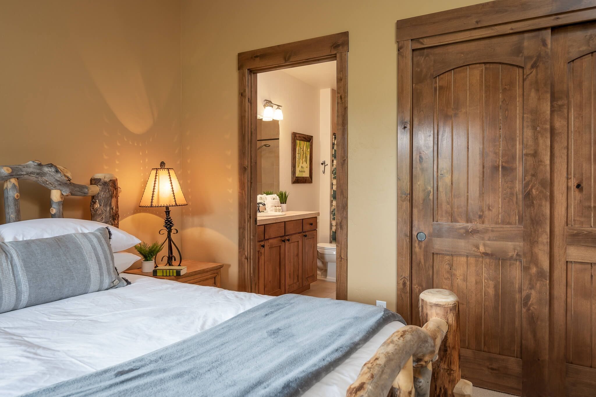 A bedroom with a wooden door, a bed with pillows, a nightstand with a lamp, and a view into the bathroom.