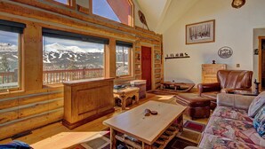 A log cabin interior with a large window offering a mountain view, a wooden desk, a leather sofa, and rustic decor including antlers and a welcome sign. - Free SkyCard Activities - Ski Area Views, Private Hot Tub - Barton Cabin (Breckenridge)