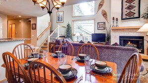 A dining area with a wooden table set for four, surrounded by wooden chairs. A staircase leads to an upper level, and a fireplace is visible in the background. - 3BR+Loft Mountain Townhome Near the Slopes. PRIVATE HOT TUB, Views! CR500 (Breckenridge)