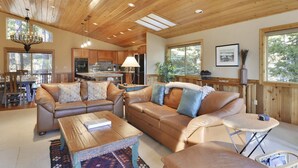 Un vaste espace de vie doté d’un plafond en bois, de canapés en cuir, d’une table basse et d’un coin repas avec des meubles de rangement en bois et un lustre.