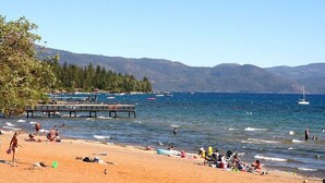 Une plage où des gens se prélassent au soleil et se baignent, une jetée qui s’avance dans l’eau et, au loin, des montagnes.