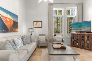 Ein Wohnzimmer mit einer Couch, Sesseln, einem Couchtisch, einem Fernseher und einem großen Fenster mit Vorhängen.