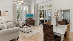 A spacious living room with a sofa, coffee table, and dining area with wicker chairs. A television is on a cabinet, and there are large windows with curtains. - Poolside Condo w/ GOLF CART! Tram to Beach. Steps to Pool and Baytowne Wharf! (Miramar Beach)