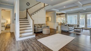 A spacious living area with a wooden staircase, two sofas, a coffee table, and a large window with blinds. - 52 N. Forest Beach-4th row (176 yard walk) to  ocean w/ elevator & much more! (Hilton Head Island)