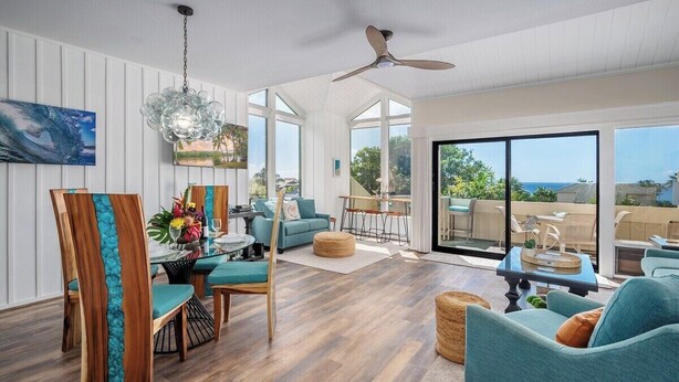 A spacious living area with a wooden dining table, teal chairs, and a large window offering a view of the ocean.