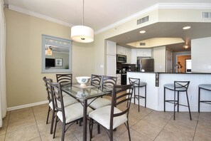 A dining area with a glass table, chairs, a hanging light fixture, and a kitchen in the background.
