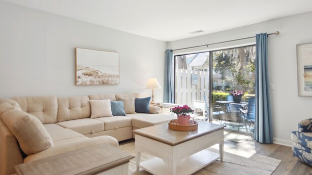 A living room with a beige sectional sofa, a white coffee table, and a large sliding glass door leading to an outdoor patio. - Courtside 67 - Short Walk to the Beach - No Steps (Hilton Head Island)