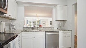 A modern kitchen with white cabinetry, a stainless steel dishwasher, and a granite countertop. - Courtside 67 - Short Walk to the Beach - No Steps (Hilton Head Island)