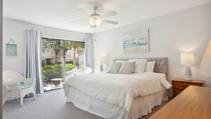 A bedroom with a large bed, a ceiling fan, a window with curtains, a chair, and a table. - Courtside 67 - Short Walk to the Beach - No Steps (Hilton Head Island)