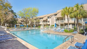 A swimming pool with lounge chairs and umbrellas, surrounded by a multi-story building with balconies. - Courtside 67 - Short Walk to the Beach - No Steps (Hilton Head Island)