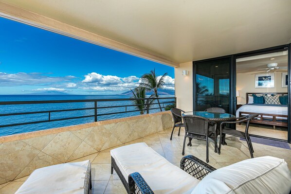 A balcony with a view of the ocean, a dining area with wicker furniture, and a bedroom with a bed and a nightstand.