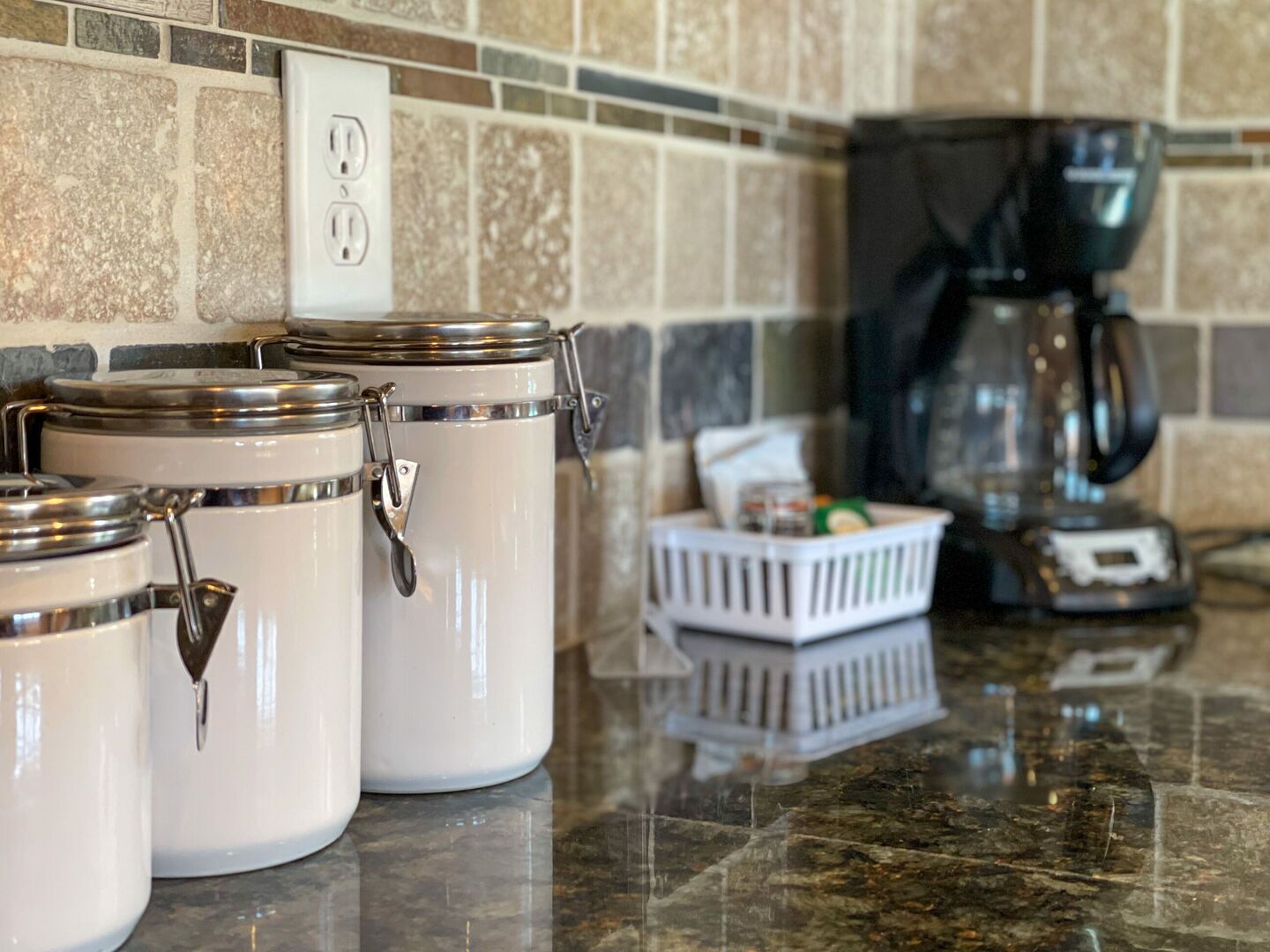 A kitchen counter with three white storage containers with metal lids and handles, a coffee maker, and a white basket on the counter.
