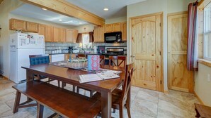 Una cocina con armarios de madera, una mesa de comedor con sillas, un refrigerador, un microondas y una ventana con cortinas.