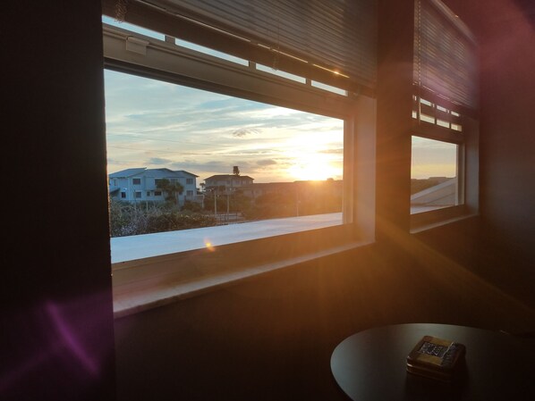 A room with a view of a sunset over a residential area, a table with a book, and a window with blinds.