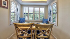 A bay window with a dining table and chairs, white shutters, and a framed picture on the wall.