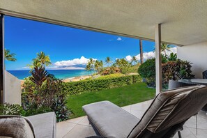 A balcony with a view of a beach and ocean, a grassy area, and a dining table.