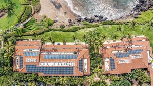 A resort with multiple buildings featuring rooftop solar panels, surrounded by lush greenery and a beach.